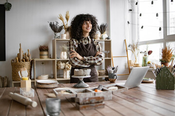 Confident Caucasian female artisan in cozy workspace surrounded by crafting materials, laptop, and...