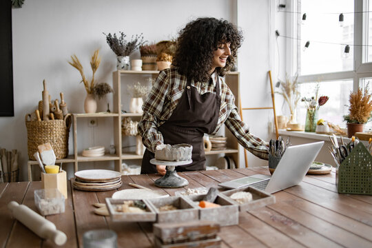 Female potter working in ceramics studio, shaping clay on pottery wheel and using laptop for research and inspiration.