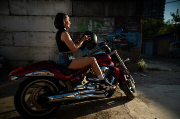 Brunette woman in denim shorts puts on a helmet while sitting on a red motorcycle. 