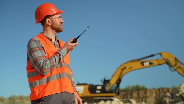 Foreman in uniform giving commands to employee in excavator using walkie talkie at construction site