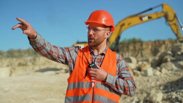 Foreman in uniform giving commands to employee in excavator using walkie talkie at construction site