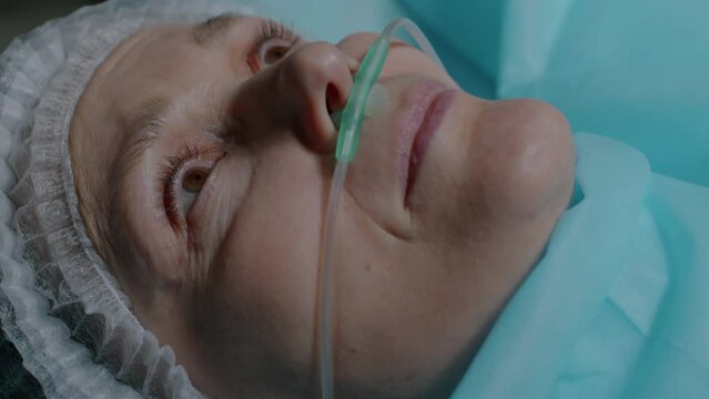From above closeup of Caucasian adult woman with oxygen tube in her nose waking up from anesthesia on operating table after surgery