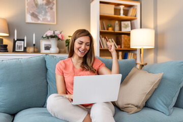 Young woman using laptop at home, looking at screen, chatting, reading or writing email, sitting on...