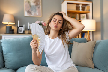 Woman puts head on sofa cushions feels sluggish due unbearable heat, waves hand fan cool herself,...