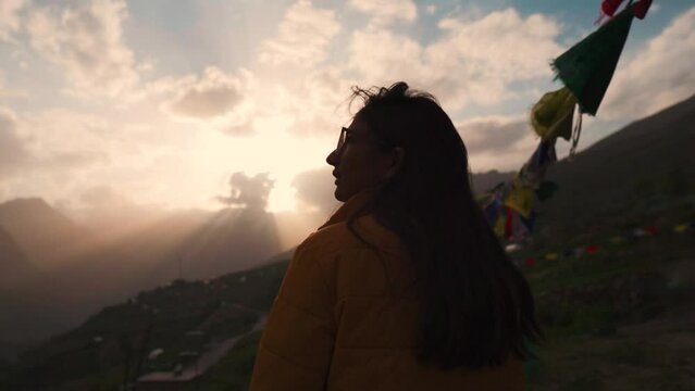 Silhouette of girl looking at orange sunset sky in mountains at Lahaul, Himachal Pradesh, India. Dramatic sunset sky with orange clouds and sunrays. Prayer flags fluttering in wind during evening. 