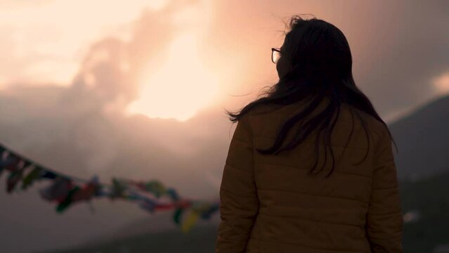 young Indian woman traveler enjoying beautiful sunset view at Himalayan mountains in Lahaul and Spiti, India. Cloudy sunset sky and mountain with Buddhist prayer flags fluttering in wind. Rack Focus
