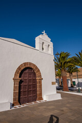 Fototapeta premium view of the main facade of the church of the village of Femés. Brown wooden door. Row of palm trees. Town square. Clear sky. Lanzarote, Canary Islands, Spain.