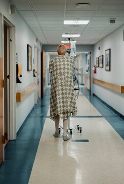 elderly male cancer patient wearing gown walks down hall in hospital