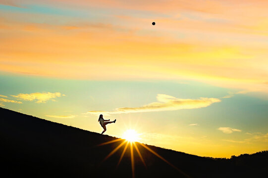 Silhouette female kicking soccer ball above sunset
