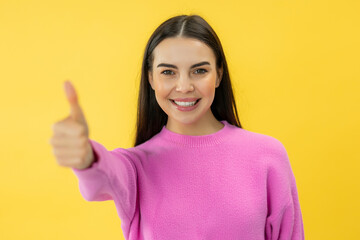 Charming happy woman with positive smile showing thumb up gesture while posing over yellow studio...