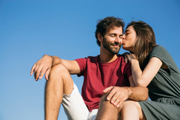 Young woman kissing man while sitting against clear sky