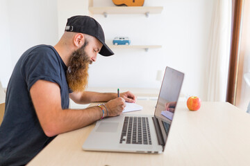 Bearded young man sitting at desk with laptop writing down something
