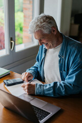 Happy senior man writing on paper while using calculator and laptop at home