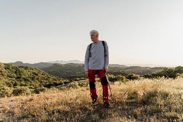 Senior male hiker looking away while standing with hiking pole on plants against clear sky