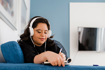 Woman with down syndrome writing in book while listening music on sofa in living room