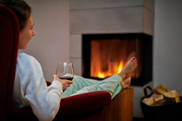Woman sitting on armchair with glass of red wine relaxing in front of fireplace