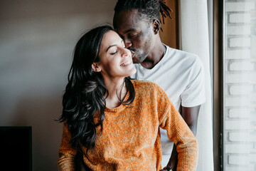 Young man kissing happy pregnant woman by window at home