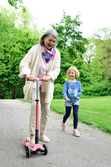 Cheerful senior woman riding push scooter while granddaughter walking in park