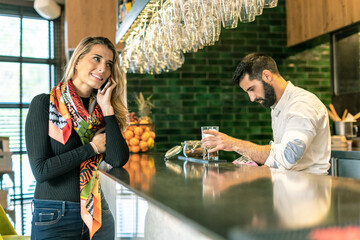Smiling woman on cell phone at the counter of a bar with barkeeper preparing a cocktail