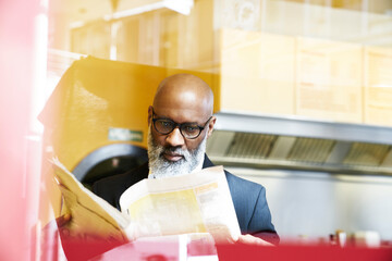 Mature businessman sitting in snack bar, reading newspaper