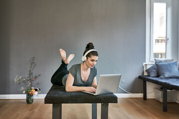 Young woman with headphones lying on lounge using laptop