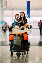 Mother holding a baby girl at the airport and pushing a trolley with the luggage