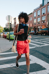 Young woman with headphones and smart phone crossong street in Brooklyn, carying take away drink