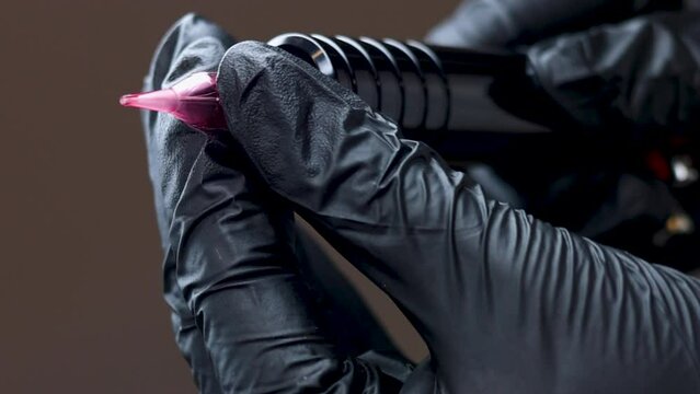 master permanent makeup attaching micro needle on tattoo machine or putting brown and red pigment in mini cup.woman in black or pink gloves, dark background.pen for micropigmentation.