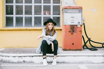 Portrait of fashionable young woman wearing hat sitting beside an old petrol pump