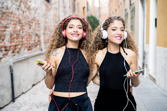 Portrait of smiling twin sisters listening music with headphones and cell phones in the city