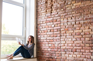 Woman sitting at home on the window sill, reading a book