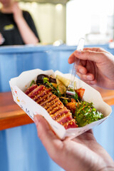 Hand holding a paper container with seared tuna and salad in a casual dining setting