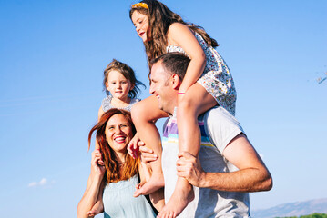 Cheerful parents carrying daughters on shoulders against clear sky during sunny day