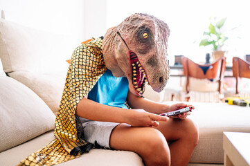 Boy wearing dinosaur mask and cape using smart phone while sitting on sofa at home