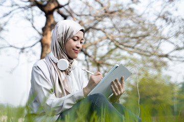 Cheerful Asian muslim woman enjoying outdoor in the morning while working on her task with tab....