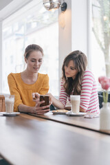 Two girlfriends meeting in a coffee shop, using smartphone