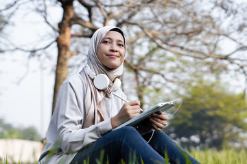 Cheerful Asian muslim woman enjoying outdoor in the morning while working on her task with tab. Student college enjoying summer outdoor while doing her task on tab