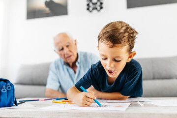 Portrait of little boy drawing with coloured pencils while his grandfather in the background watching him