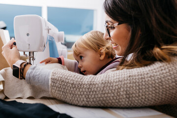 Happy mother with little daughter at home using sewing machine