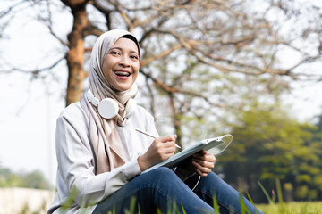 Cheerful Asian muslim woman enjoying outdoor in the morning while working on her task with tab. Student college enjoying summer outdoor while doing her task on tab