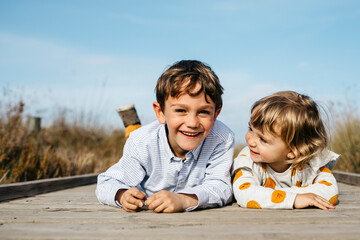 Portrait of boy and his little sister lying side by side on boardwalk having fun