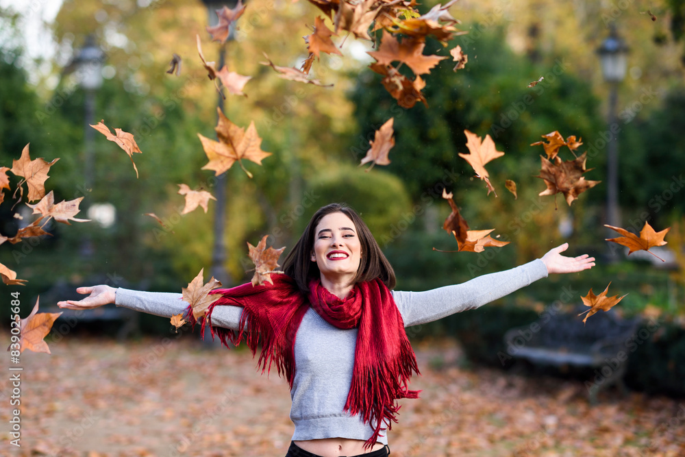 Wall mural portrait of laughing young woman with red scarf throwing autumn leaves in the air