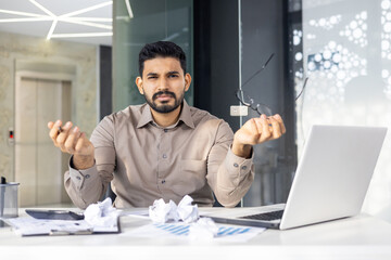 Frustrated businessman holding eyeglasses with a puzzled expression at office desk, surrounded by...