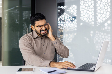 A businessman sitting at his desk, experiencing ear pain and discomfort while working on a laptop. Office environment suggests stress or health issues during work.