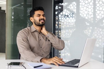Man in an office setting, sitting at a desk, holding his neck in pain while using a laptop,...