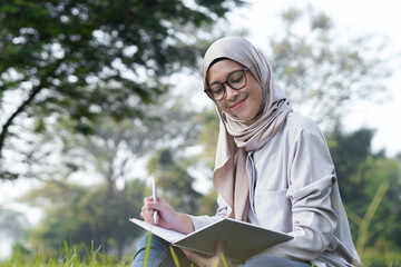 A joyful woman sits cross-legged in a green park, holding a notebook, captured from a low left angle. She looks at her notebook with a smiling and thoughtful expression, enjoying the peaceful outdoor