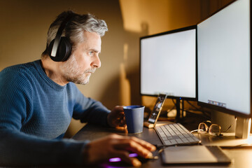 Mature man with headphones sitting at desk at home working on computer