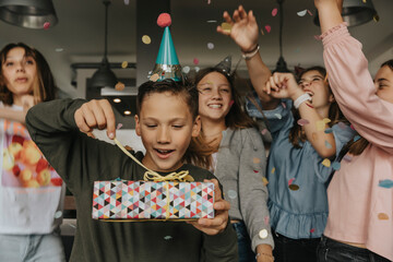 Curious birthday boy opening gift while friends dancing in background