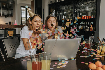 Cheerful friends blowing confetti while video conferencing over laptop while sitting at home