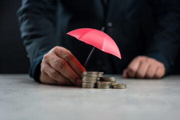 Close up businessman hand holding a red umbrella over stack of coins on table to protect saving....